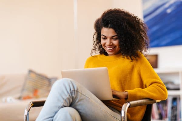 Image of cheerful african american woman using laptop while sitting on chair in living room